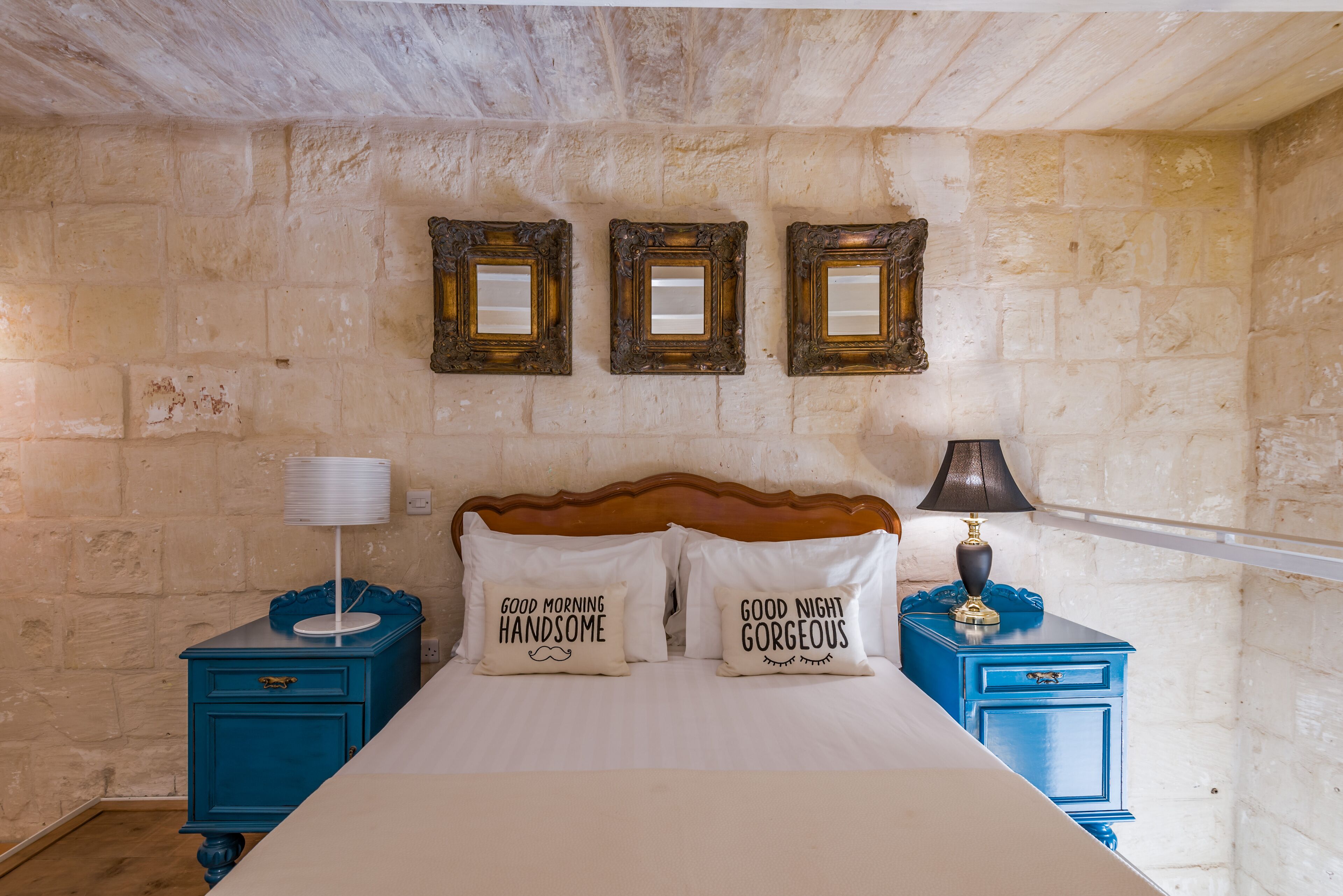A bedroom with a stone wall, a wooden headboard, two bedside tables with lamps, and two pillows with text.