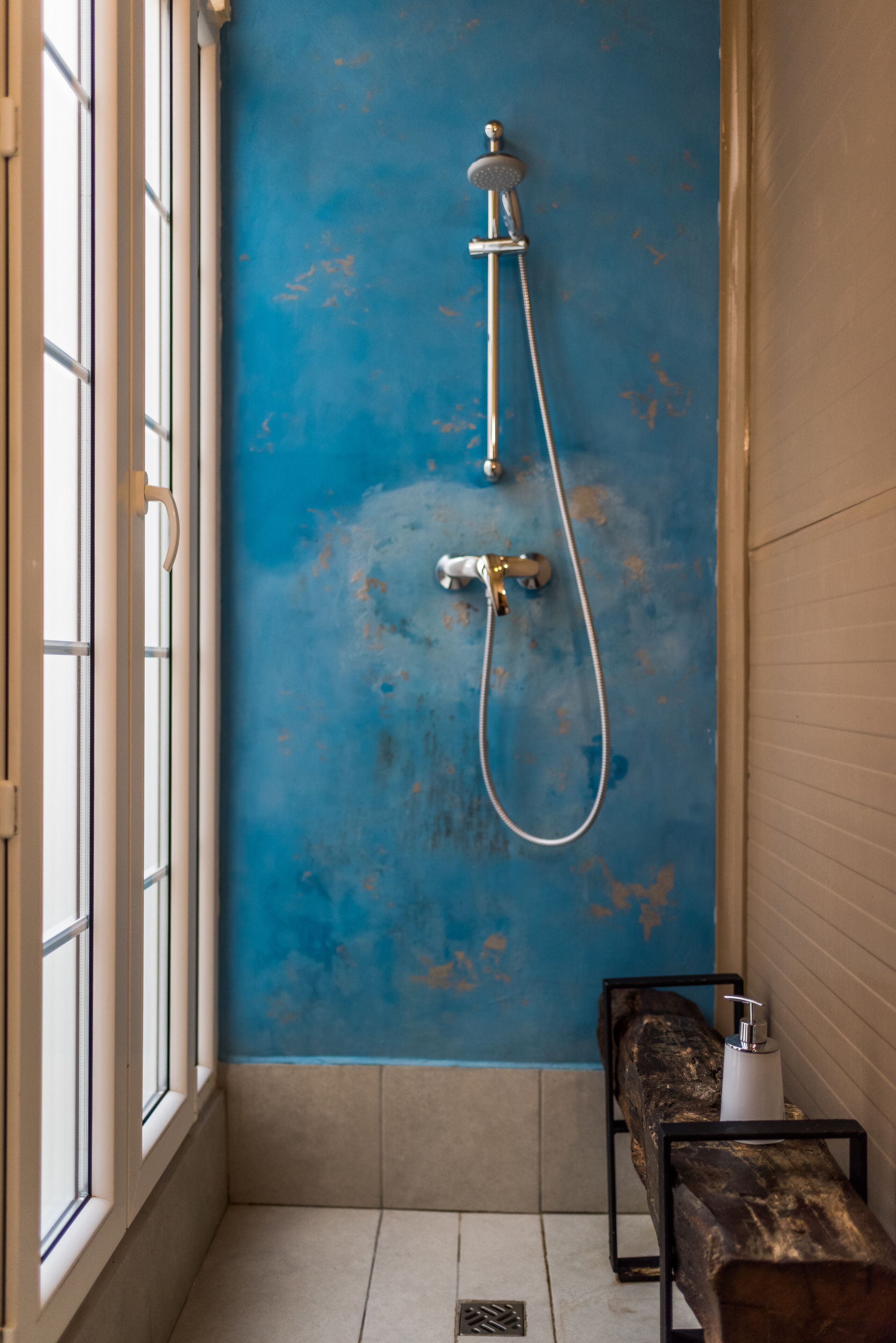 A bathroom with a blue wall, a shower, a bench, and a window.