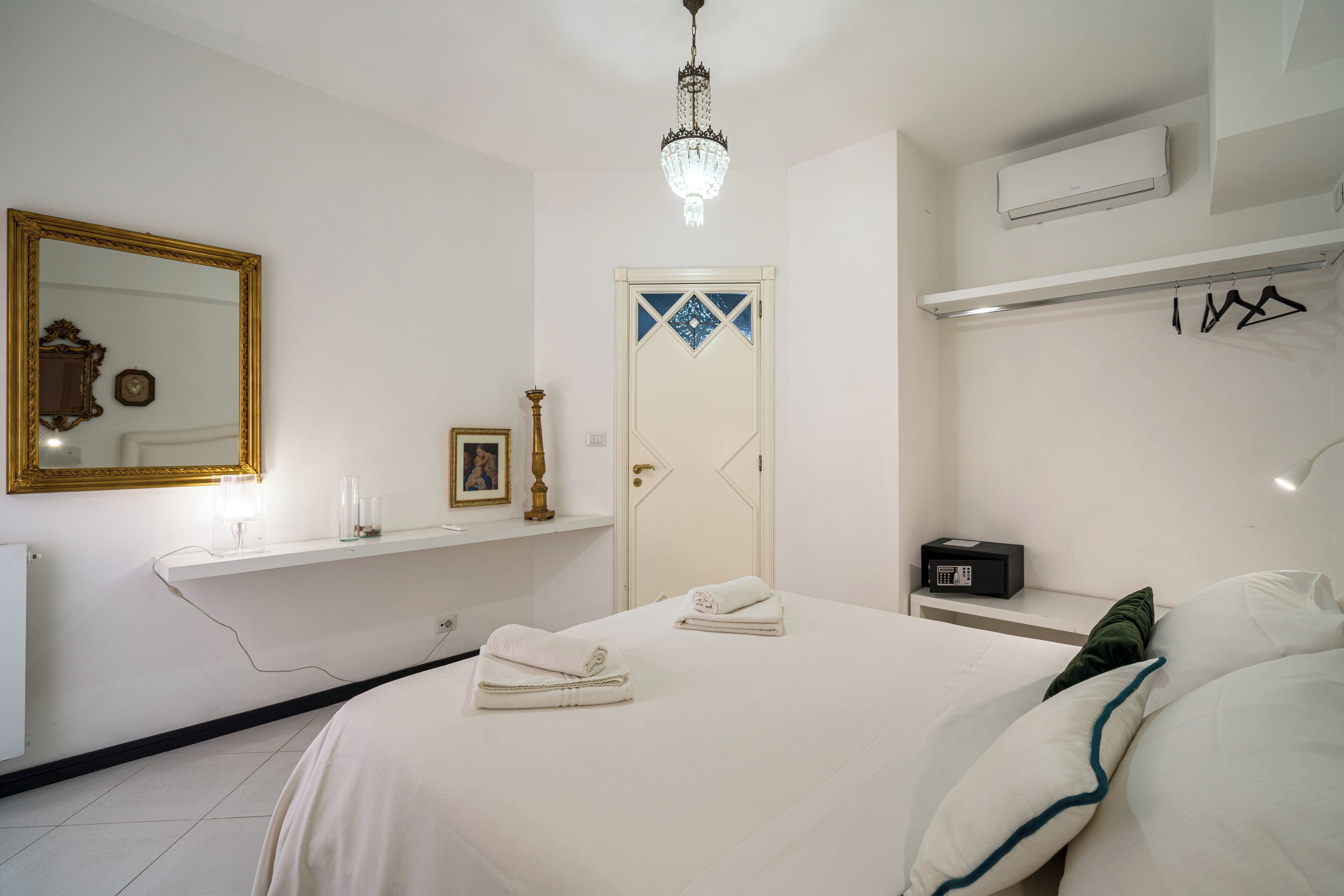 A hotel room with a bed, a bedside table with a radio, a mirror, a chandelier, and a door with a decorative design.