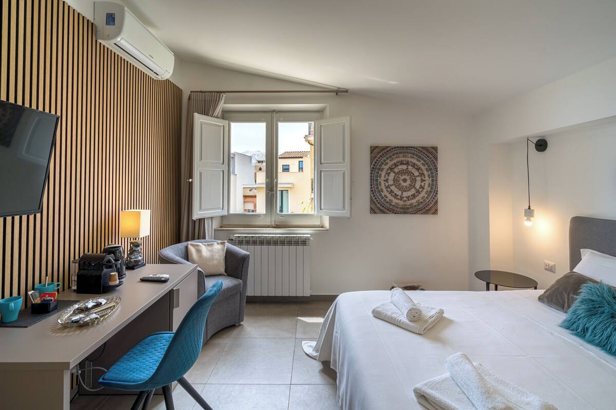 A hotel room with a bed, a desk, a chair, a TV, a window, and a radiator.