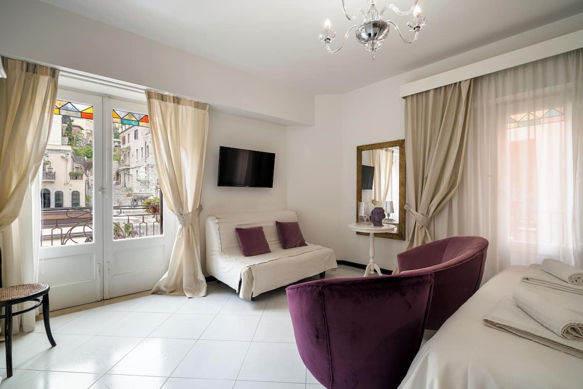 A hotel room with a bed, a purple armchair, a small table, a TV, and a window with curtains.