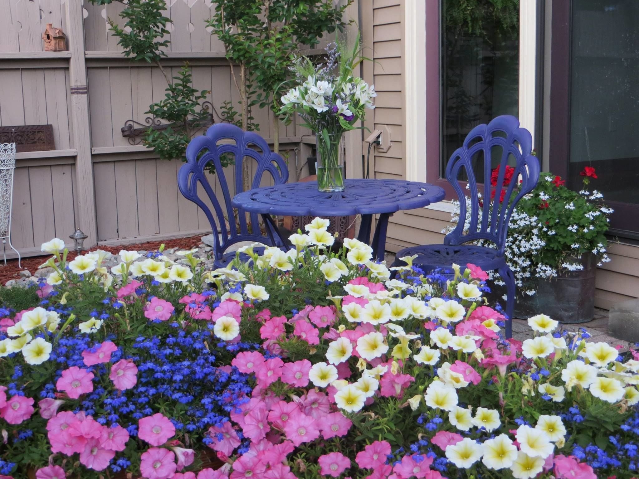 A patio with a round table and two chairs, surrounded by a variety of colorful flowers.