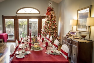 A dining room with a red table set for a meal, a Christmas tree, and a cabinet with a lamp.