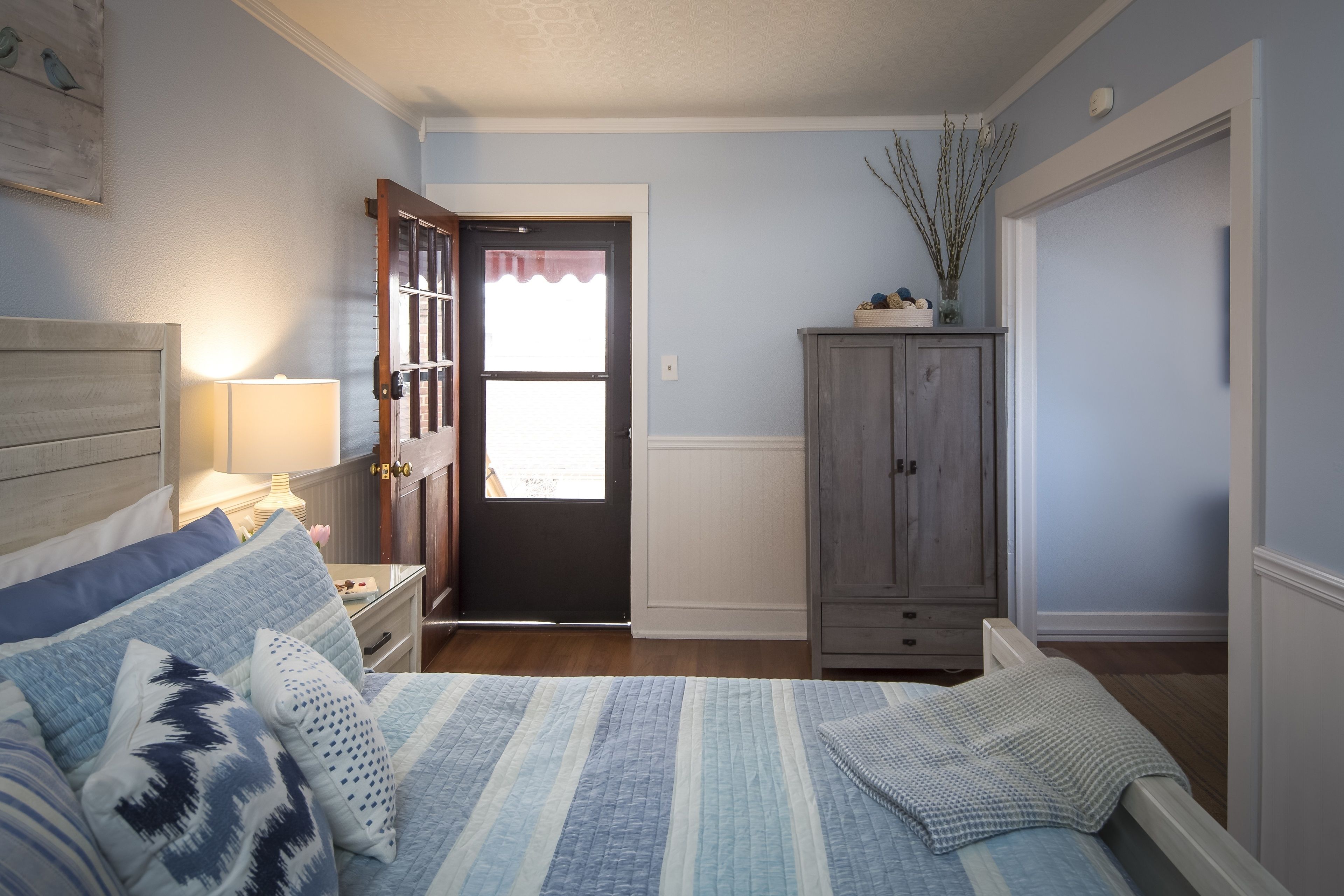 A bedroom with a bed, nightstand, lamp, dresser, and a door leading to another room.