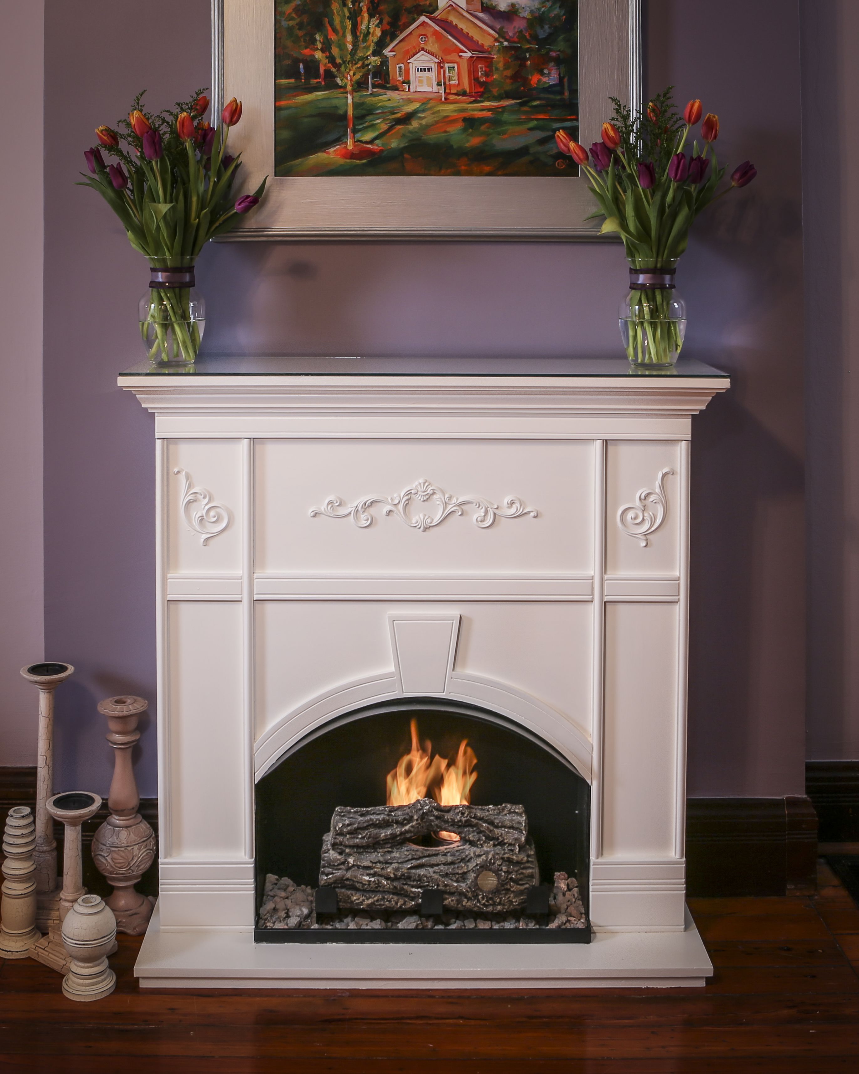 A white fireplace with a fire, flanked by two vases of tulips, and a framed painting above.