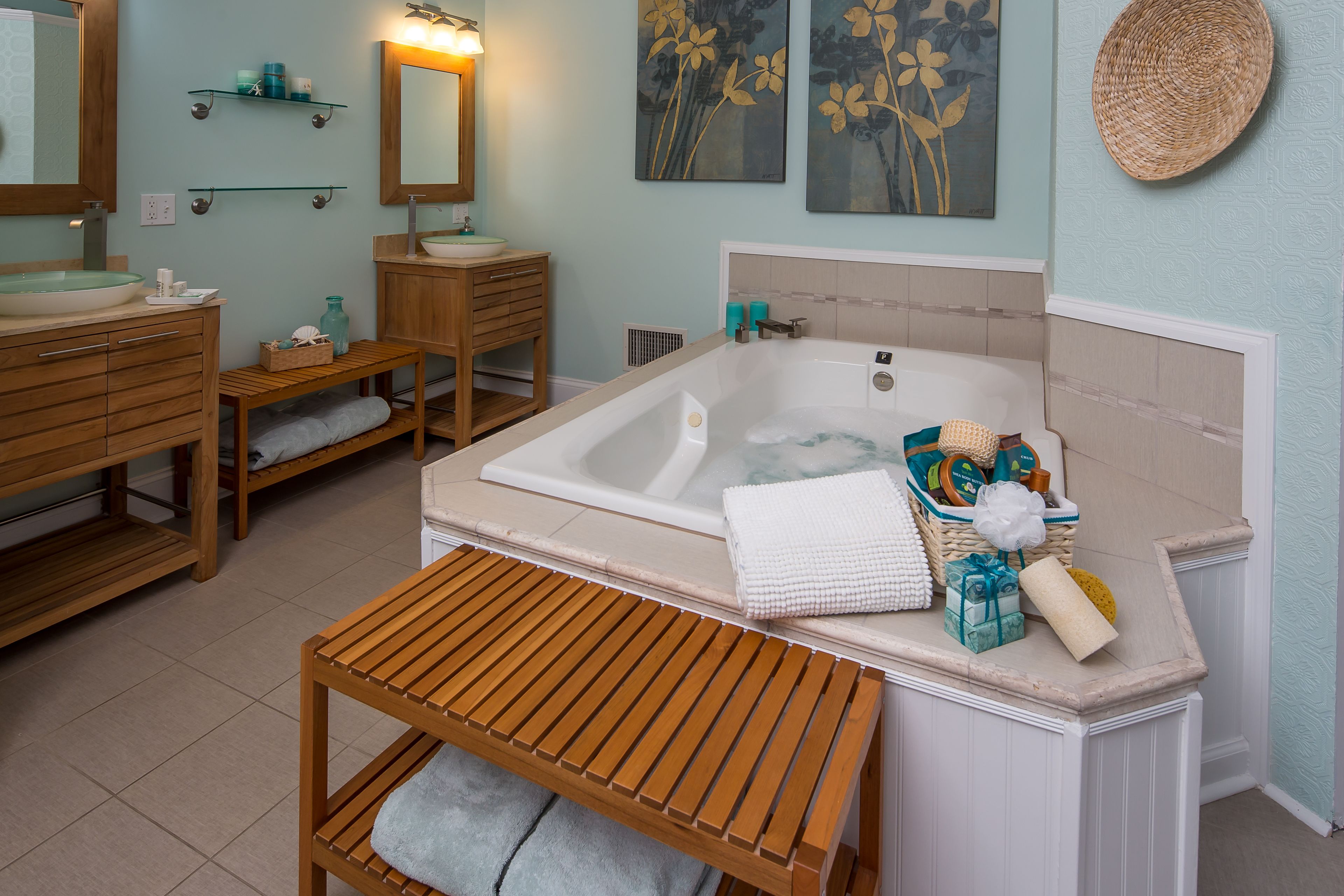 A bathroom with a bathtub, wooden bench, shelves with toiletries, and artwork on the walls.