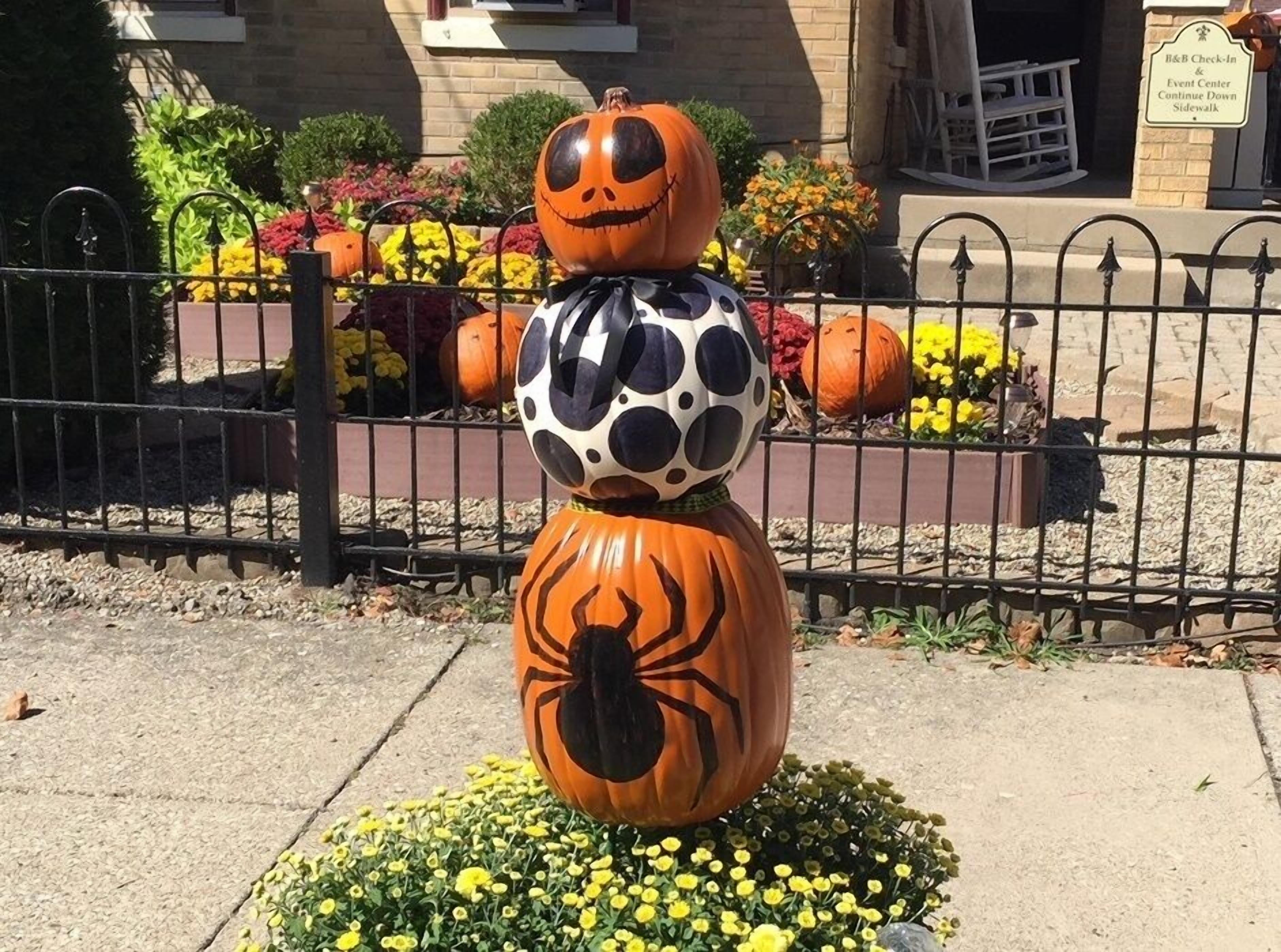 A pumpkin decoration with a spider design in front of a fence with flowers and a building in the background.