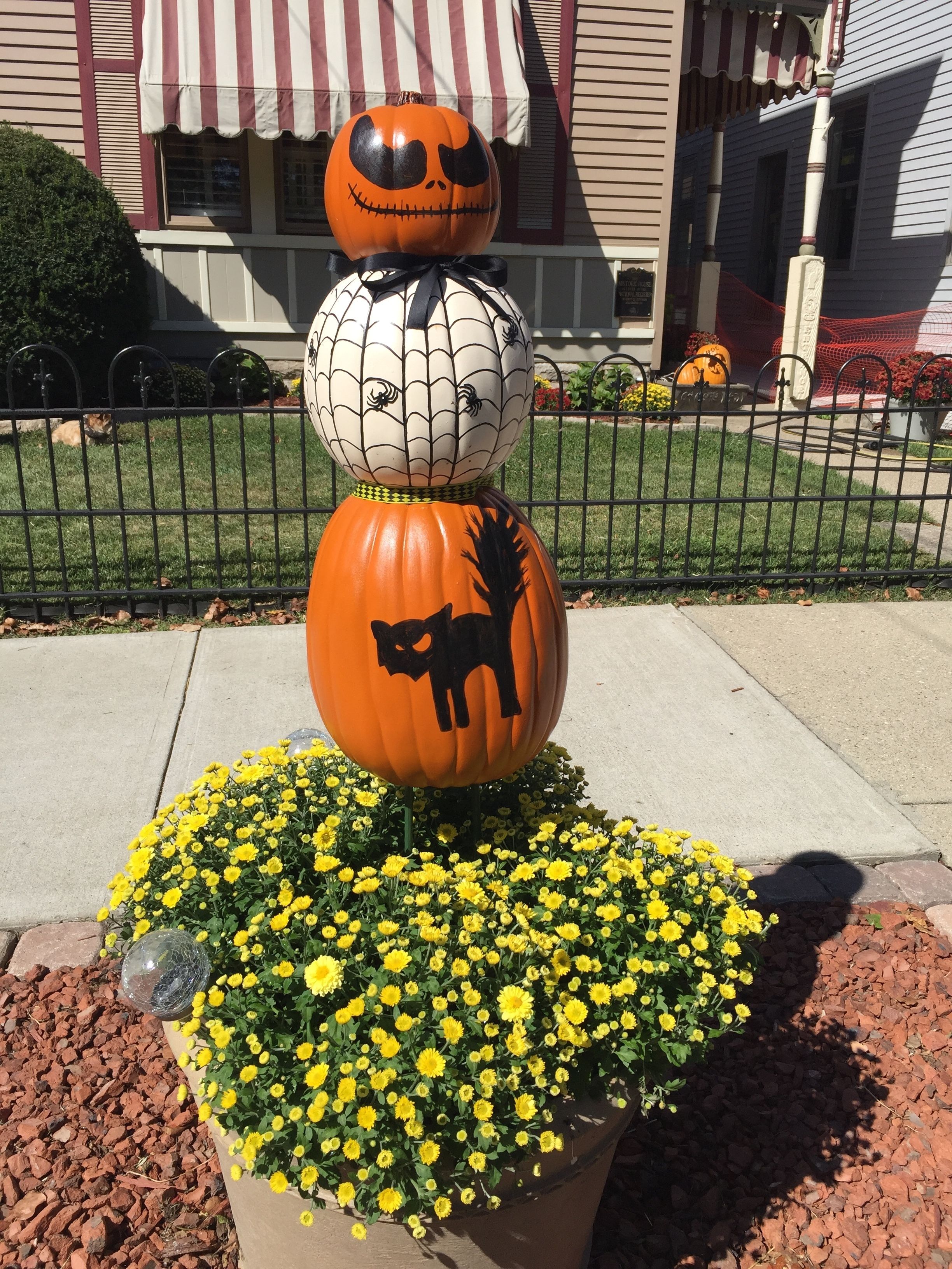 Un arreglo decorativo de calabazas con una tela de araña y la silueta de un gato, frente a un edificio con toldo a rayas.