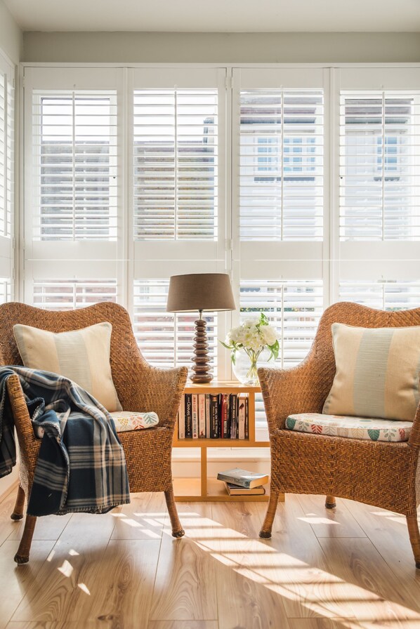 A room with two wicker armchairs, a small wooden table with a lamp, a bookshelf, and a window with blinds. - Beachcomber - an apartment that sleeps 4 guests in 2 bedrooms (Rye)