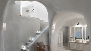 A modern interior with a white staircase, a wooden chair, a large rock, and a bathroom with a sink and mirror. - Villa Ioli Anastasia (Santorini)