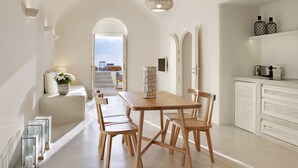 A modern kitchen with a wooden dining table, white cabinetry, and a view to the outside. - Villa Ioli Anastasia (Santorini)