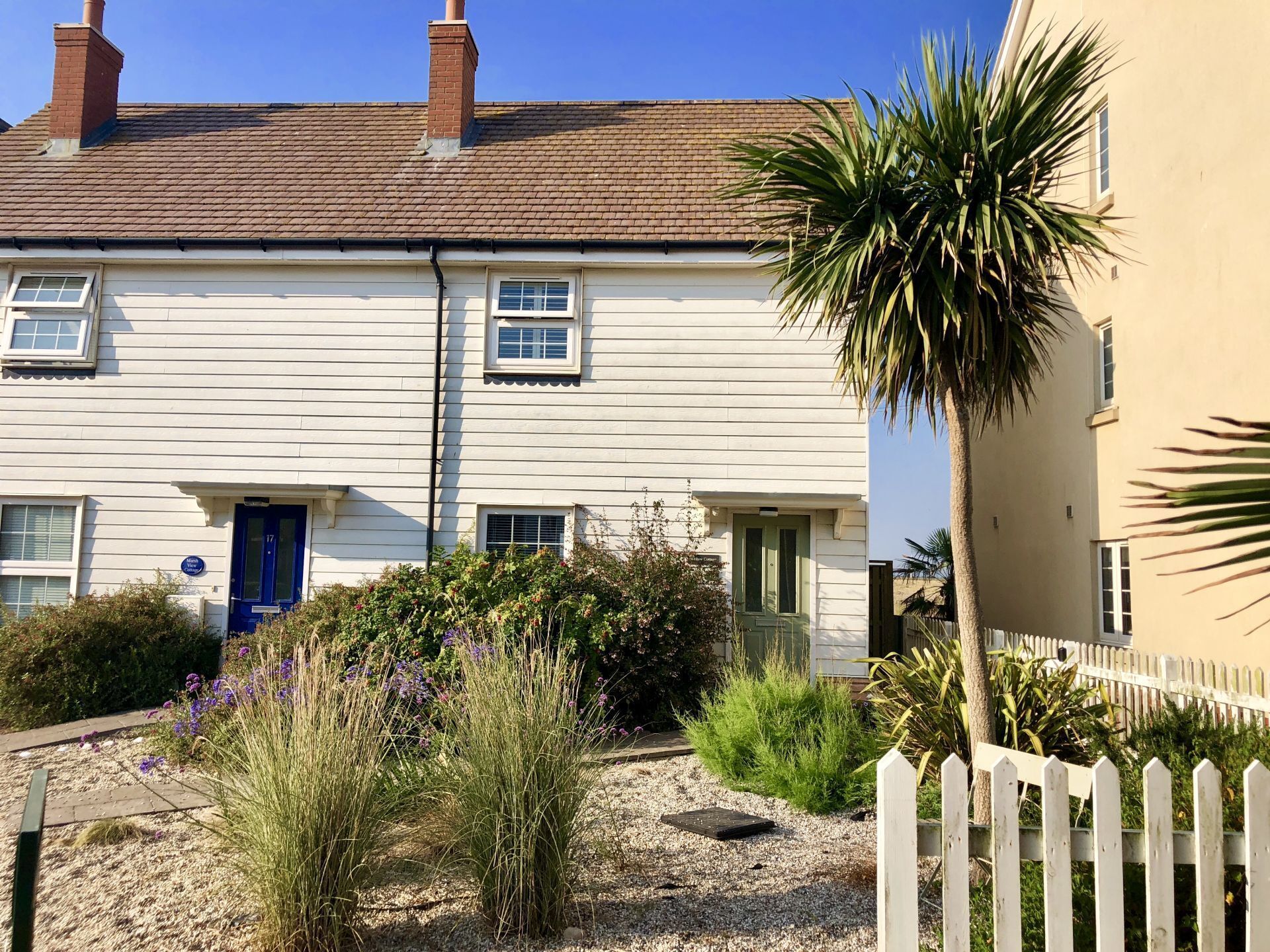 A two-story house with a blue door, a small garden with various plants, and a white picket fence.