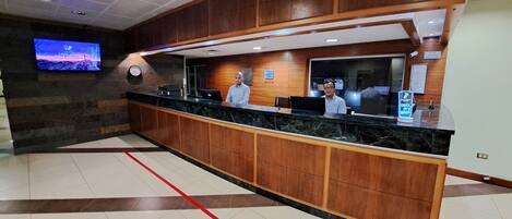 A reception desk with a digital screen displaying a cityscape, wooden paneling, and a clock on the wall.