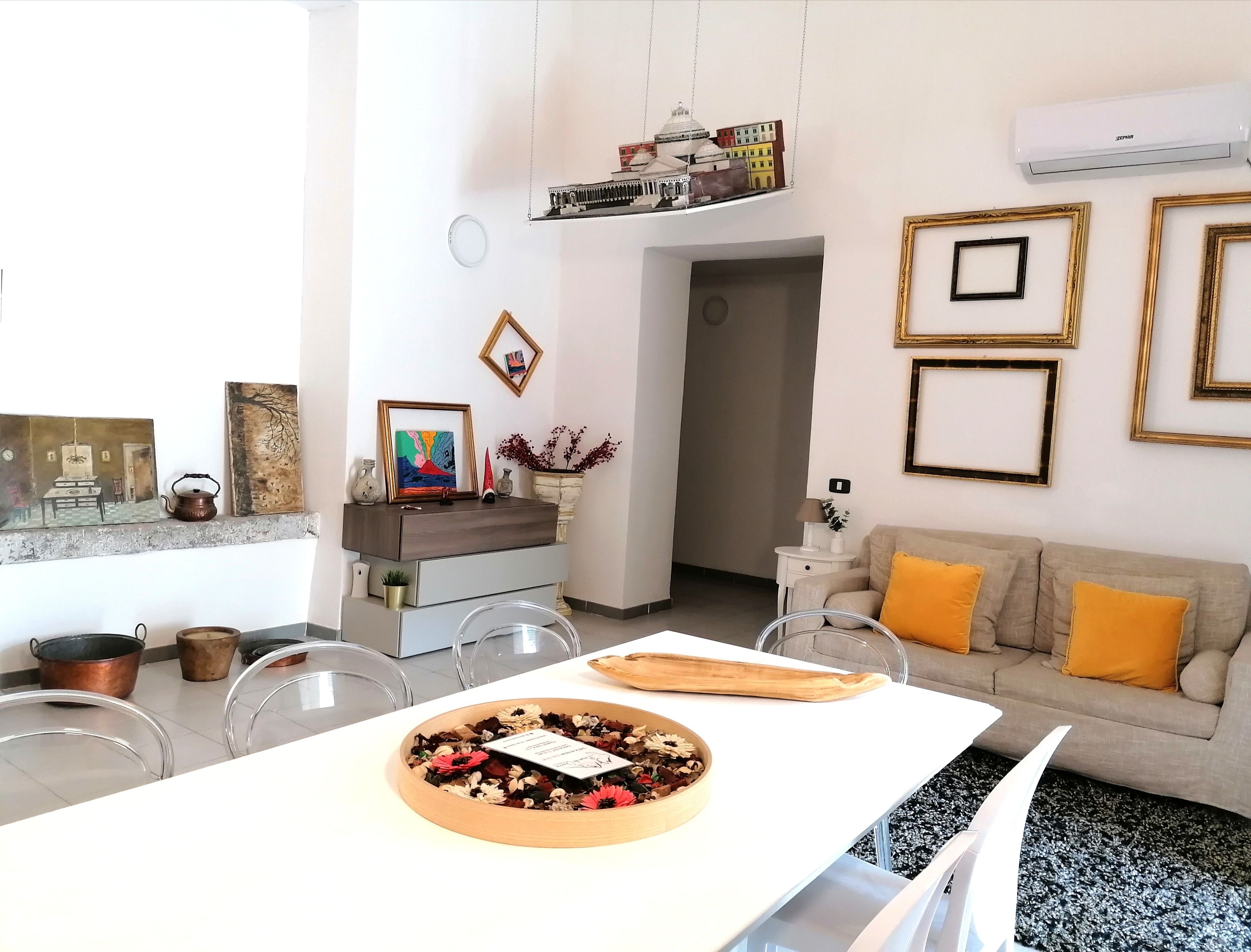 Una sala de estar moderna con un sofá blanco, una mesa de comedor con un centro de mesa floral y una zona de cocina con utensilios de cobre.