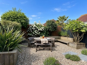 A patio area with a round wooden table and benches, surrounded by gravel and various plants.