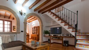 A spacious living room with wooden flooring, a sofa, a coffee table, and a TV. There is a staircase with a decorative railing and a dining area with a wooden cabinet and chairs.