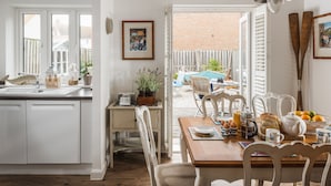 Una cocina con comedor, armarios blancos, una mesa de madera, sillas, una planta en maceta y vista al patio trasero.