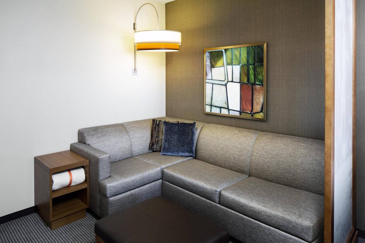 A modern hotel room with a grey sectional sofa, a small wooden nightstand with a towel, a wall-mounted light fixture, and a colorful abstract painting.