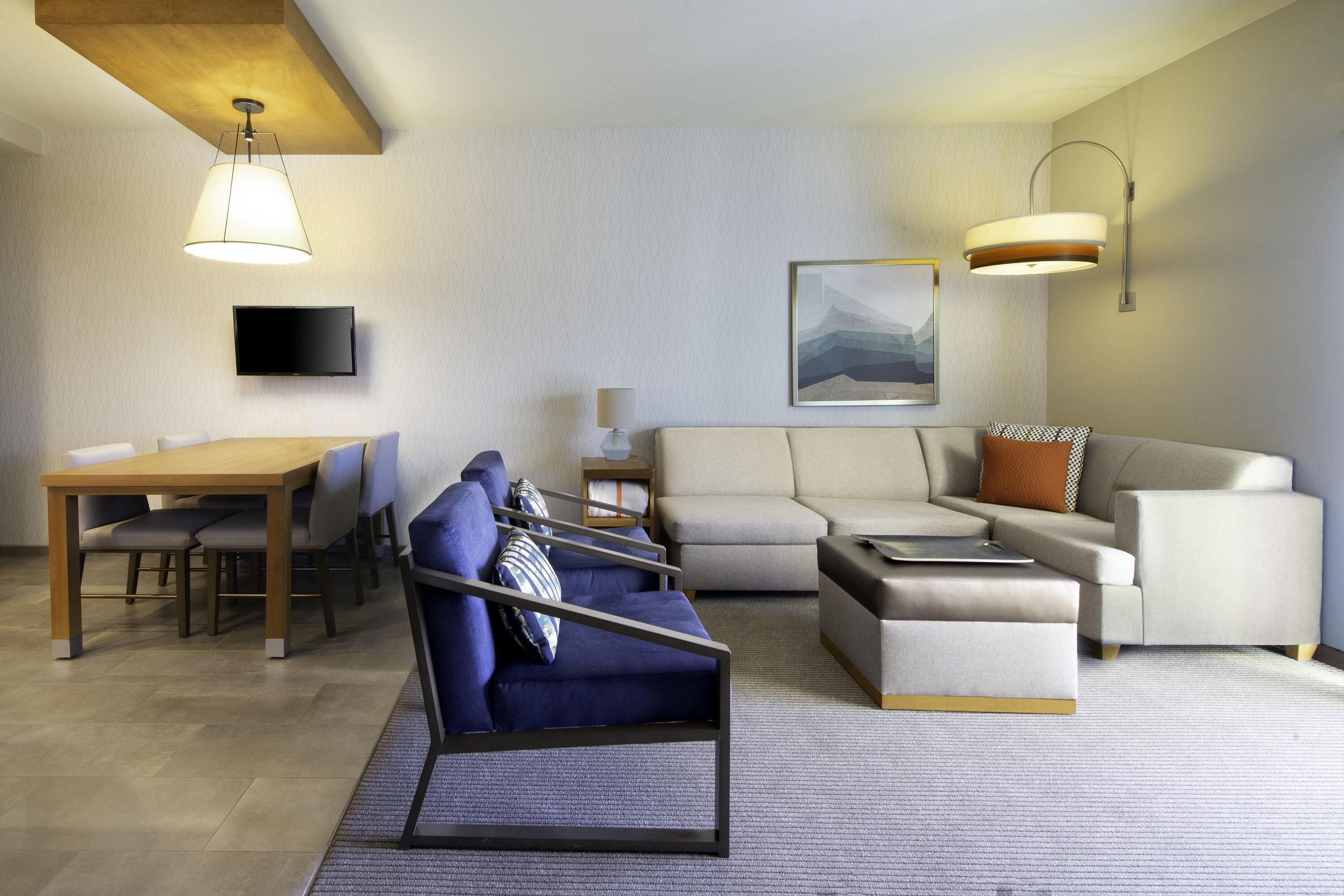A modern living room with a sofa, armchair, ottoman, dining table, chairs, a flat-screen TV, and a painting on the wall.