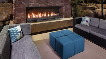 A stone fireplace with a fire, a gray sofa with a patterned pillow, and a blue ottoman in an outdoor seating area.