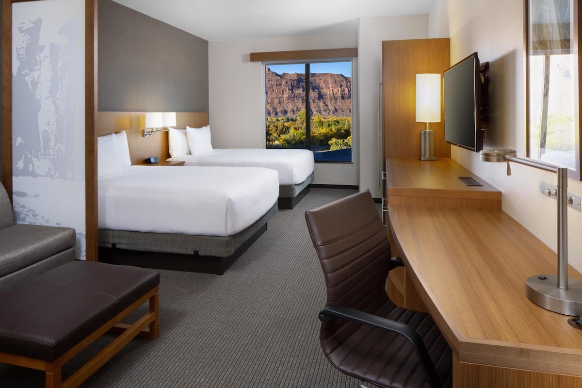 A hotel room with two beds, a desk, a TV, and a window with a mountain view.