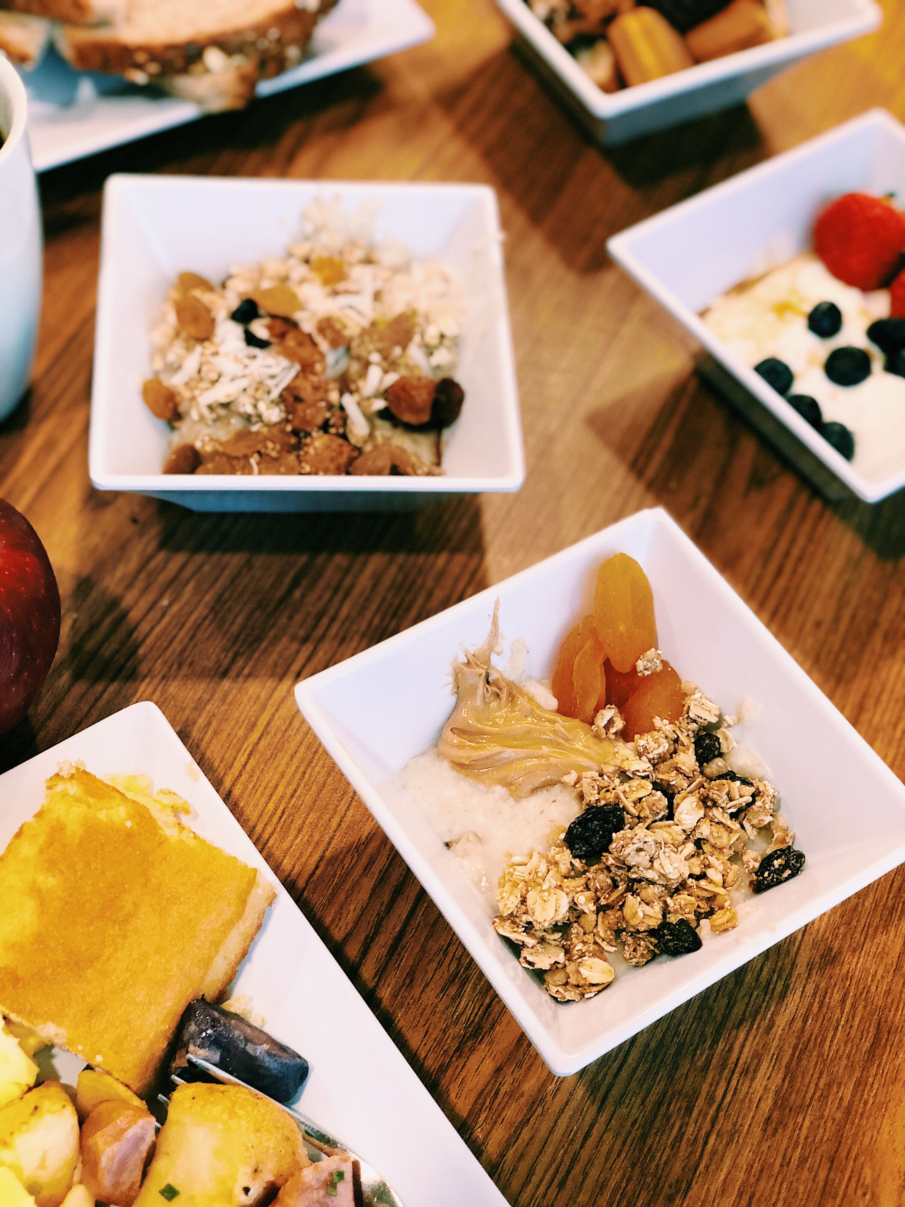 A table with various breakfast dishes in square white plates, including cereal, fruits, and a slice of cake.