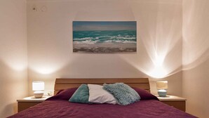 Un dormitorio con cama, dos lámparas de noche, un cuadro del océano y una cortina translúcida.