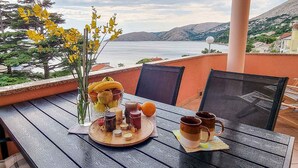 Una mesa con ajuar de frutas, velas y tazas en un balcón con vistas a un cuerpo de agua y colinas.