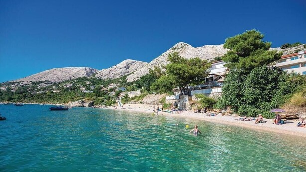 Una playa costera con agua turquesa cristalina, montañas rocosas al fondo y algunos edificios con balcones con vistas al mar.
