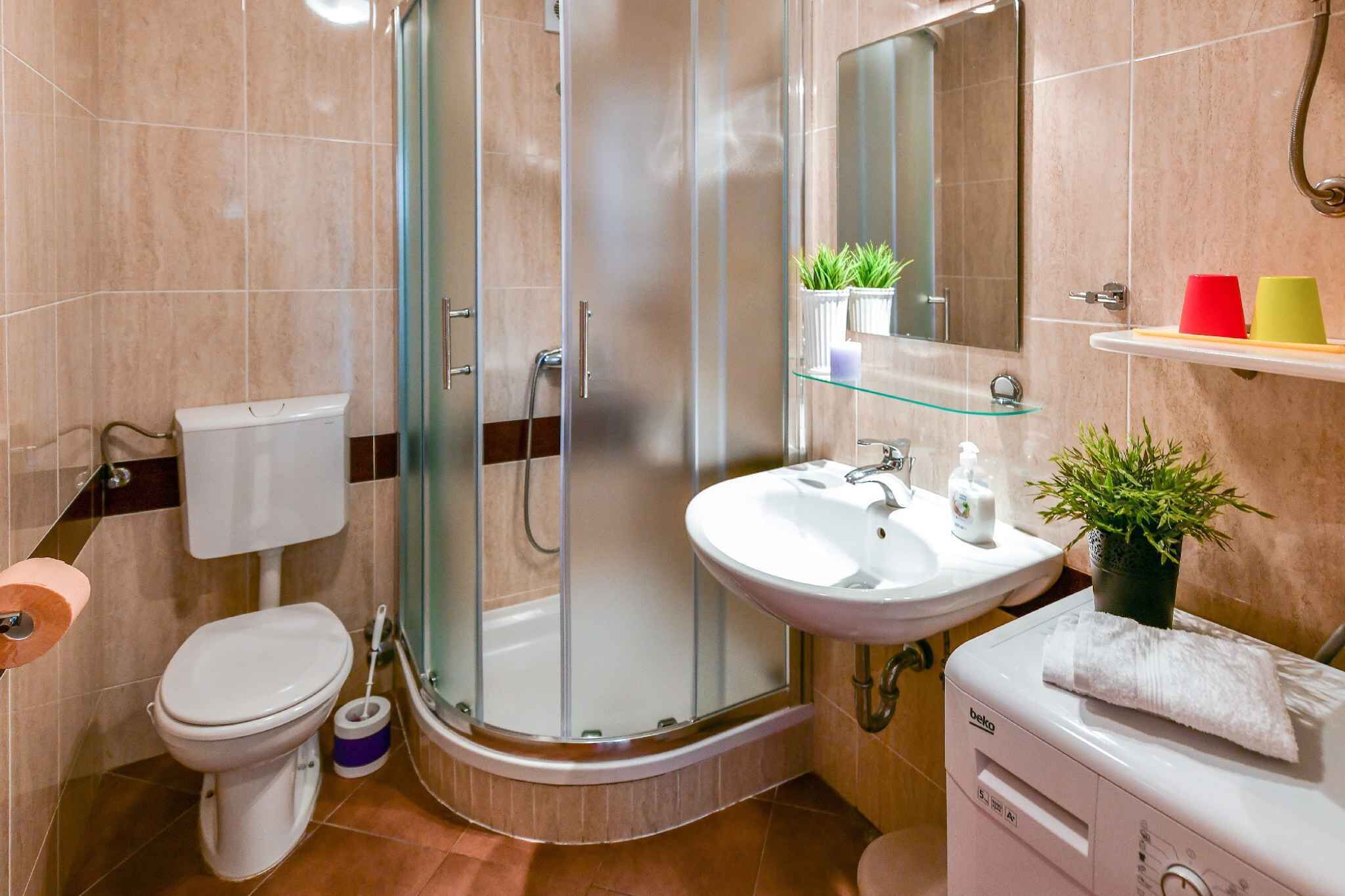 Um banheiro moderno com box de vidro, vaso sanitário branco, pia com espelho e pequeno armário com produtos de higiene pessoal.