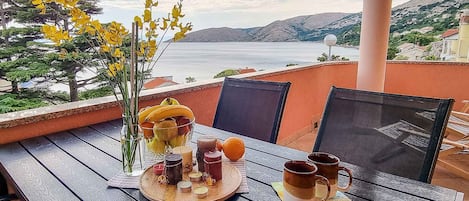A table set with a fruit basket, candles, and mugs on a balcony overlooking a body of water and hills.