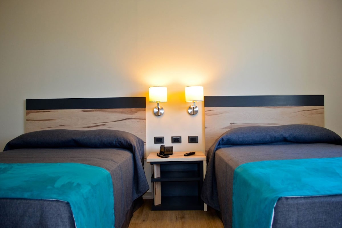 A hotel room with two beds, each with a teal bedspread, a wooden headboard, and a nightstand with a lamp and phone.