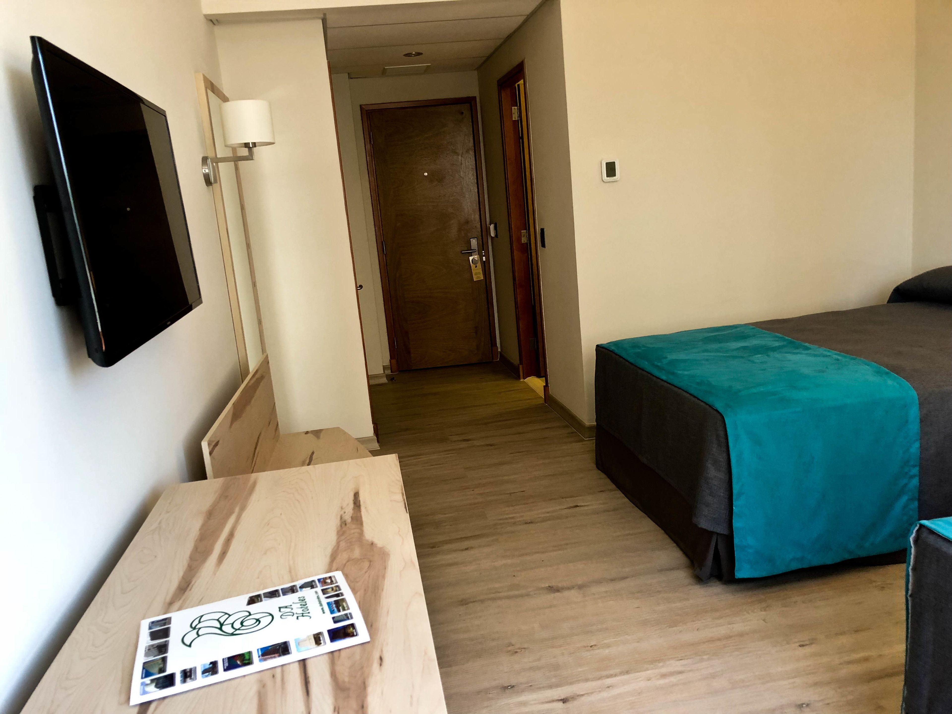 Habitación de hotel con cama, televisión montada en la pared, piso de madera y puerta que da a otra habitación.