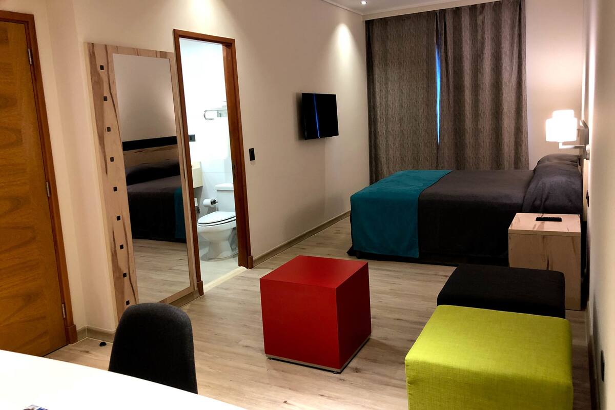 A hotel room with a bed, a TV, a mirror, a desk, and a bathroom visible through an open door.