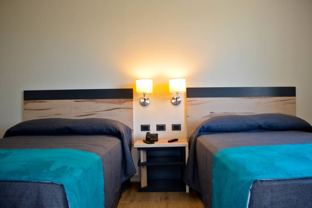 A hotel room with two beds, each with a teal bedspread, a wooden headboard, and a nightstand with a phone and remote control.