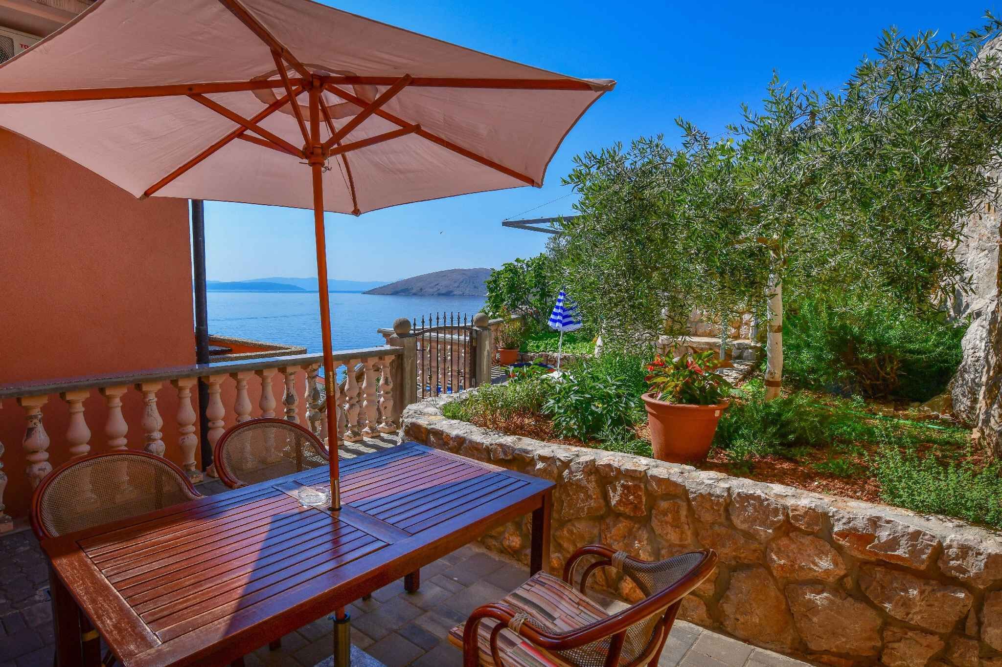 A wooden table and chairs under a large umbrella on a balcony with a stone wall and potted plants.