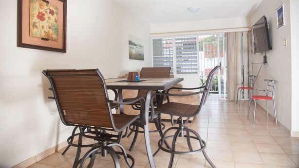 A dining area with a table and four chairs, a framed artwork on the wall, and a television mounted on the wall. - Montones Beach Apartment C-3 (Isabela)