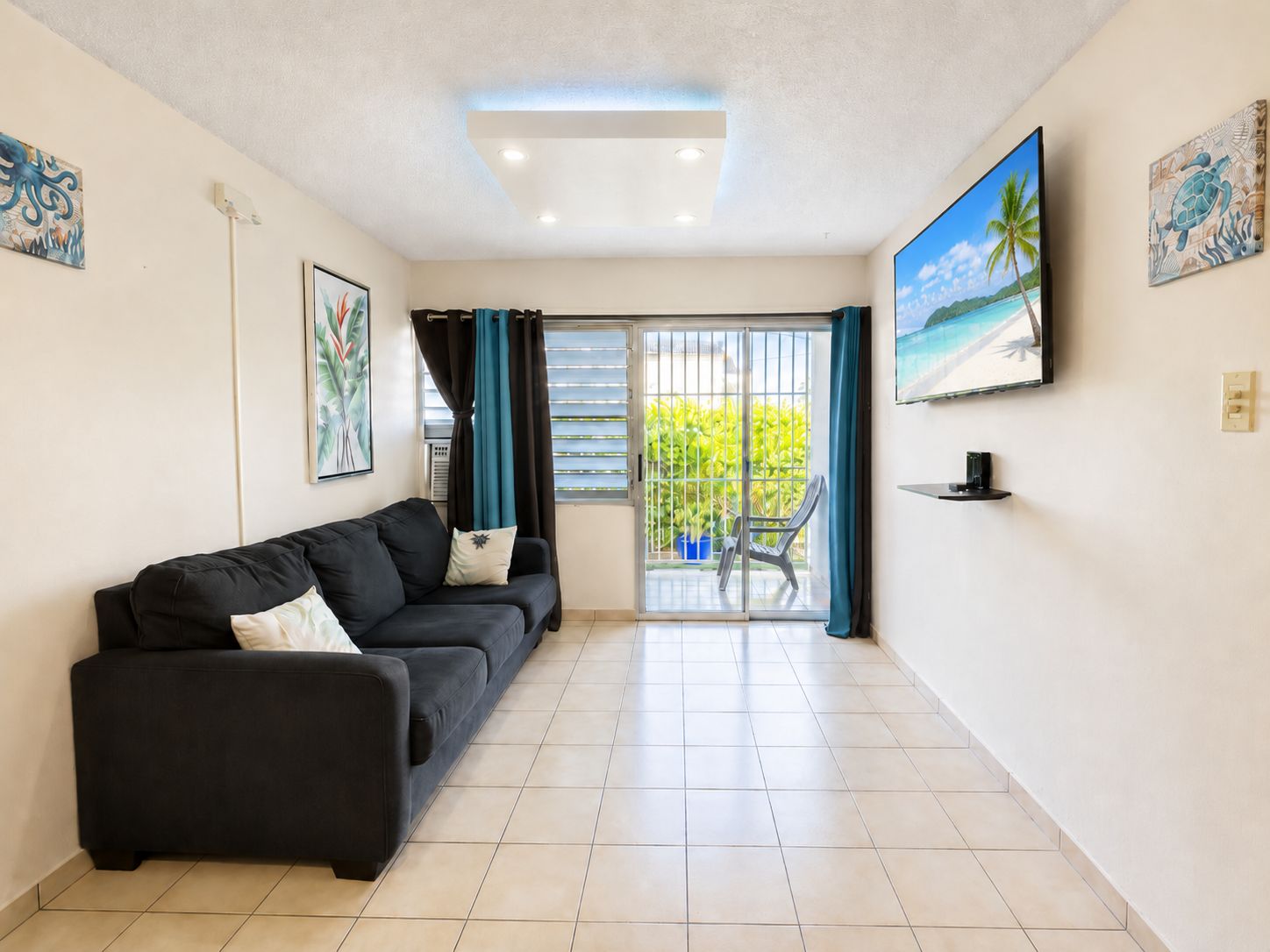 A living room with a black sofa, a sliding glass door leading to a balcony, and a TV mounted on the wall.