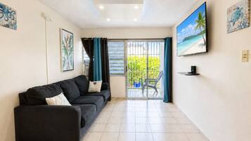 A living room with a black sofa, a sliding glass door leading to a balcony, and a TV mounted on the wall.