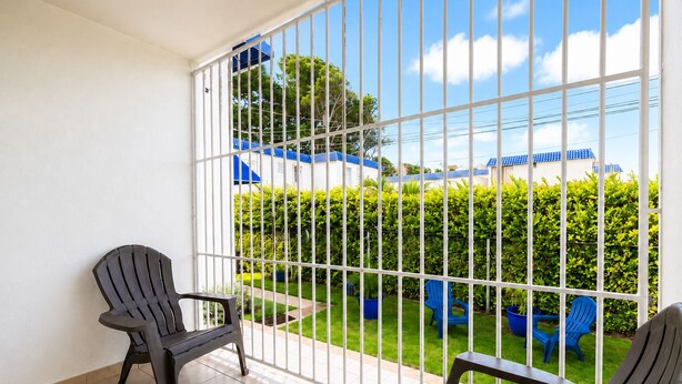A balcony with two chairs, a tiled floor, and a view of a blue-roofed building and green hedges. - Montones Beach Apartment C-3 (Isabela)