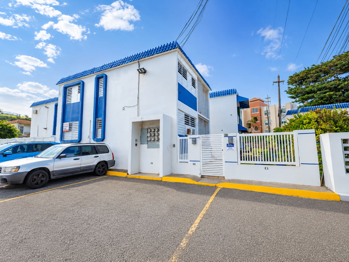 Um edifício com portão fechado, esquema de cores branco e azul, e um carro branco estacionado em frente.