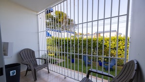 A balcony with two chairs, a trash bin, and a view of a garden and buildings. - Montones Beach Apartment C-3 (Isabela)