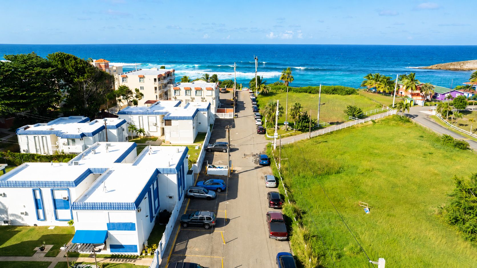 A coastal area with a row of white buildings with blue trim, a parking lot with several cars, and a grassy field with a fence.