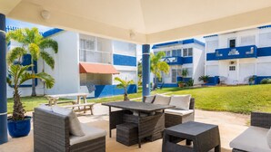 An outdoor patio area with wicker furniture, a barbecue grill, and a view of a blue and white building. - Montones Beach Apartment C-3 (Isabela)