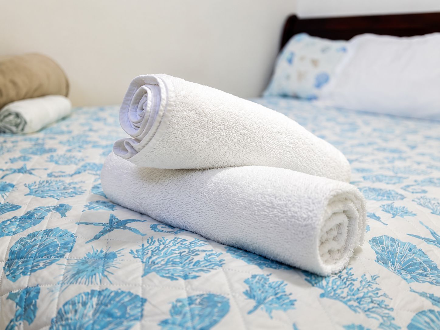 Ein ordentlich bezogenes Bett mit einem weißen, aufgerollten Handtuch, das auf einer Bettdecke mit blauem Blumenmuster liegt.