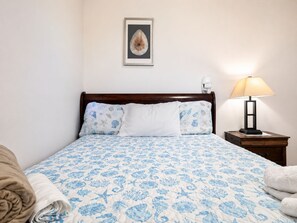 Un dormitorio con cama, almohadas, una mesita de noche, una lámpara y un cuadro en la pared.