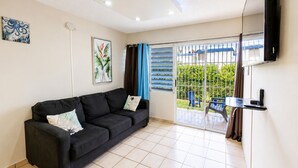 A living room with a black sofa, a flat-screen TV mounted on the wall, and a sliding glass door leading to an outdoor area with a view of greenery. - Montones Beach Apartment C-3 (Isabela)