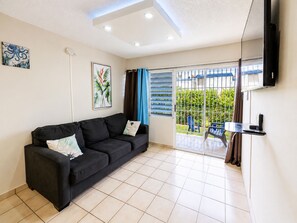 Una sala de estar con un sofá negro, un televisor de pantalla plana montado en la pared y un ventanal corredero que da a un espacio exterior con vistas a zonas verdes.