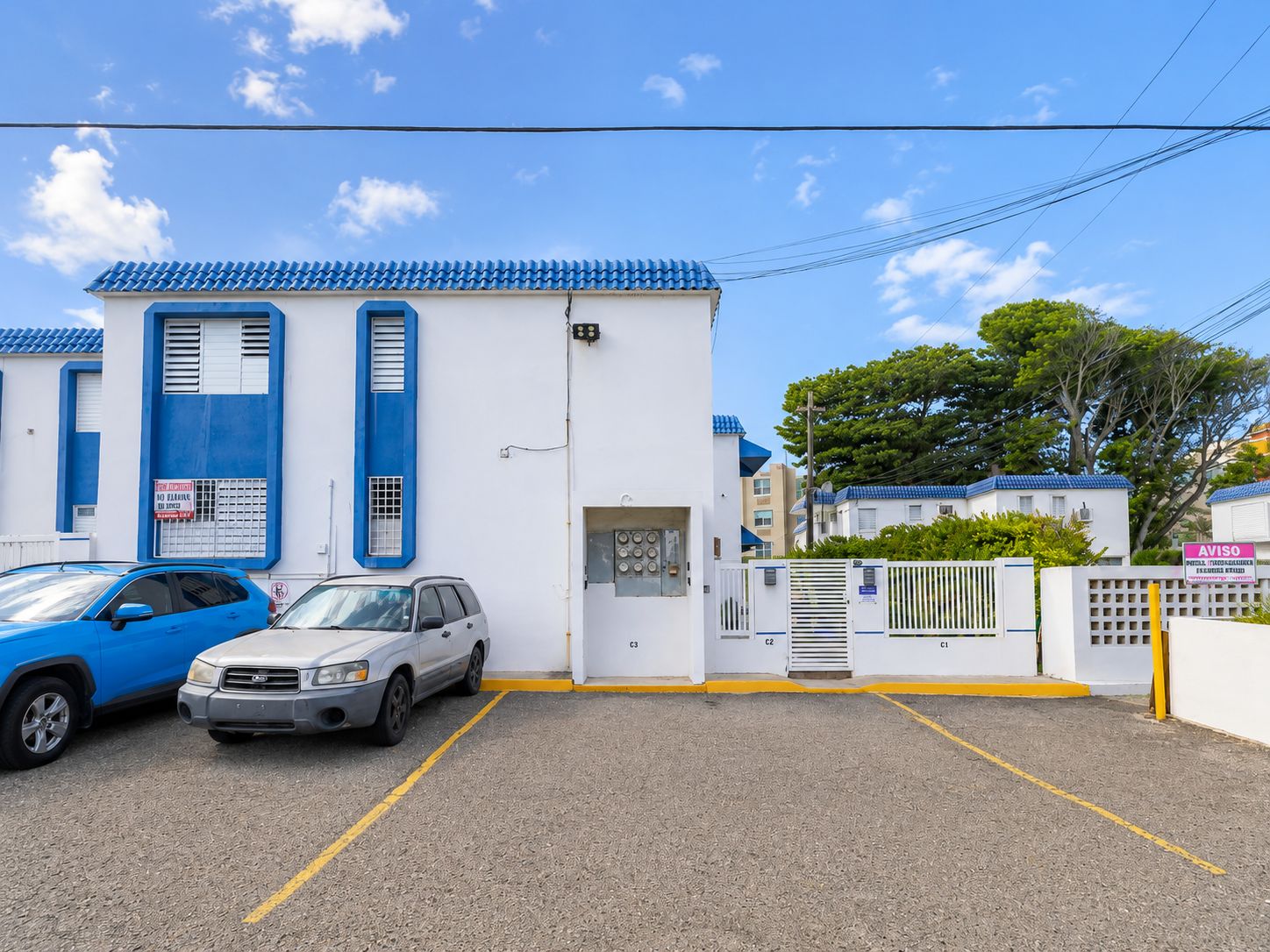 Un aparcament amb dos cotxes, un edifici blanc amb detalls blaus i una entrada tancada amb reixeta.