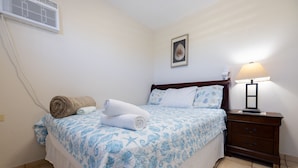 A neatly made bed with a blue floral bedspread, a wooden nightstand with a lamp, and a wall-mounted air conditioner. - Montones Beach Apartment C-3 (Isabela)